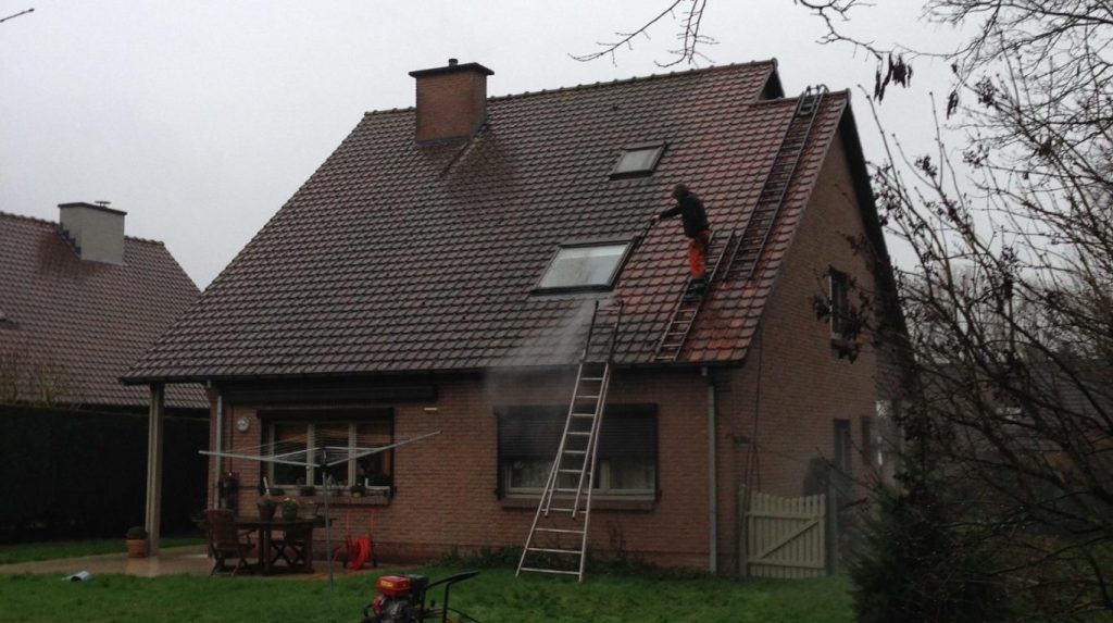 Une personne nettoie à haute pression le toit d'une maison en briques par temps nuageux, debout près du sommet avec un tuyau tandis qu'une échelle repose contre le toit.